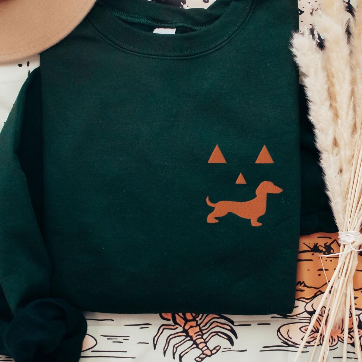 Dachshund Jack-o'-Lantern Sweatshirt – Forest Green Forest Green crewneck sweatshirt laid flat with a felt hat and dried pampas grass, embroidered on the left chest with an orange dachshund jack-o'-lantern face: triangle eyes and nose over a doxie silhouette grin