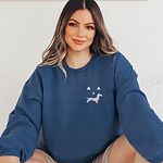 Woman sitting in a Navy crewneck sweatshirt embroidered on the left chest with a white dachshund jack-o'-lantern face