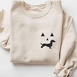 Sand crewneck sweatshirt folded on white, embroidered on the left chest with a black jack-o'-lantern face made of triangle eyes, a triangle nose and a dachshund silhouette