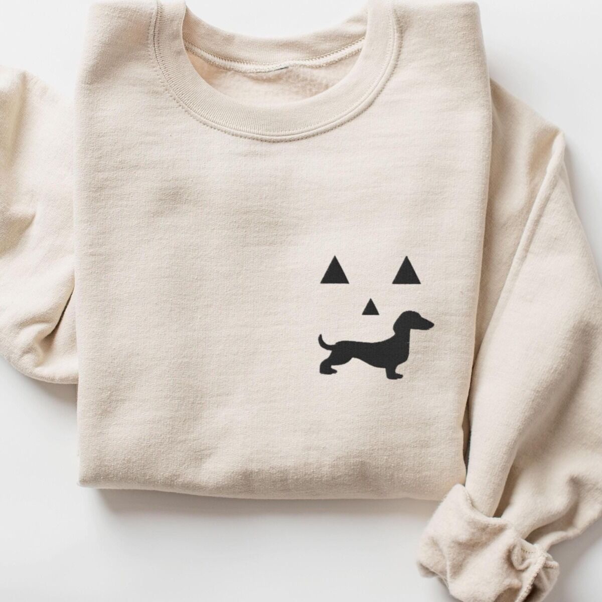Dachshund Jack-o'-Lantern Sweatshirt – Sand Sand crewneck sweatshirt folded on white, embroidered on the left chest with a black jack-o'-lantern face made of triangle eyes, a triangle nose and a dachshund silhouette