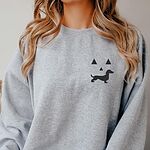 Woman in a Sport Grey crewneck sweatshirt embroidered on the left chest with a black jack-o'-lantern face whose grin is a dachshund silhouette