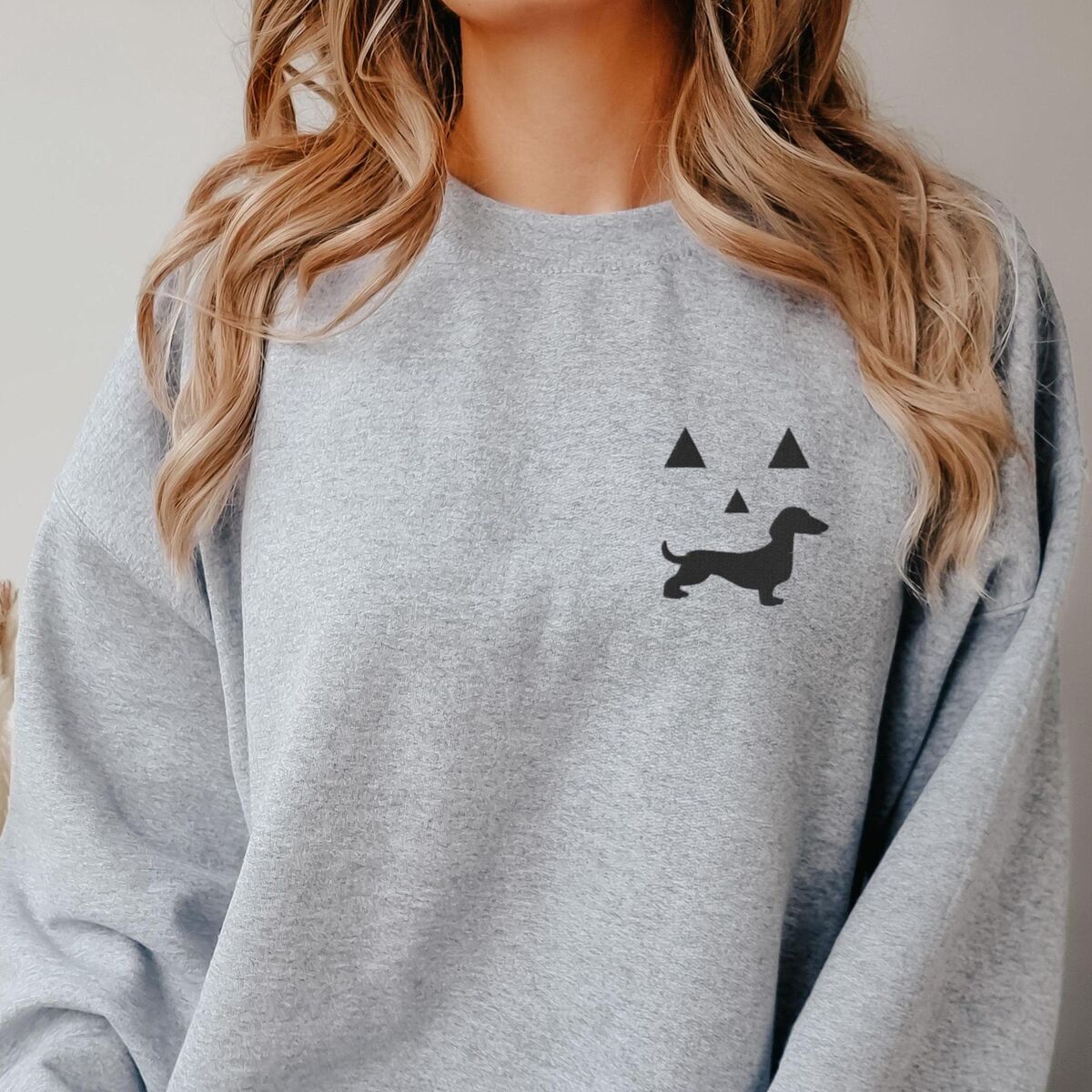 Dachshund Jack-o'-Lantern Sweatshirt - Sport Grey Woman in a Sport Grey crewneck sweatshirt embroidered on the left chest with a black jack-o'-lantern face whose grin is a dachshund silhouette