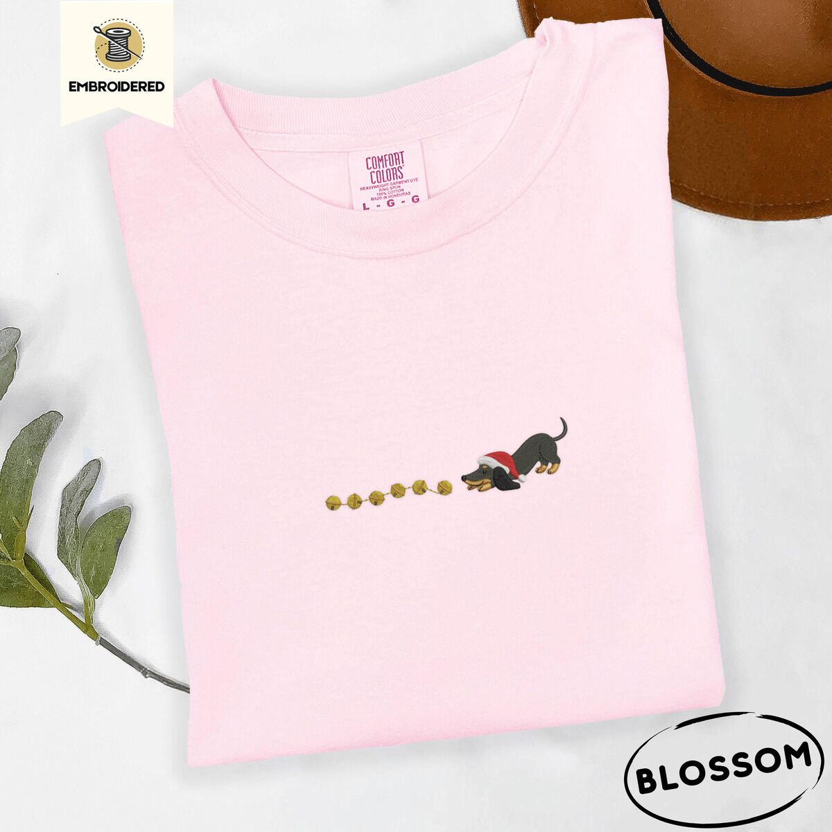 Dachshund Jingle Bells Shirt – Blossom Blossom Comfort Colors T-shirt embroidered with a black and tan dachshund in a Santa hat bowing down to sniff a string of gold jingle bells