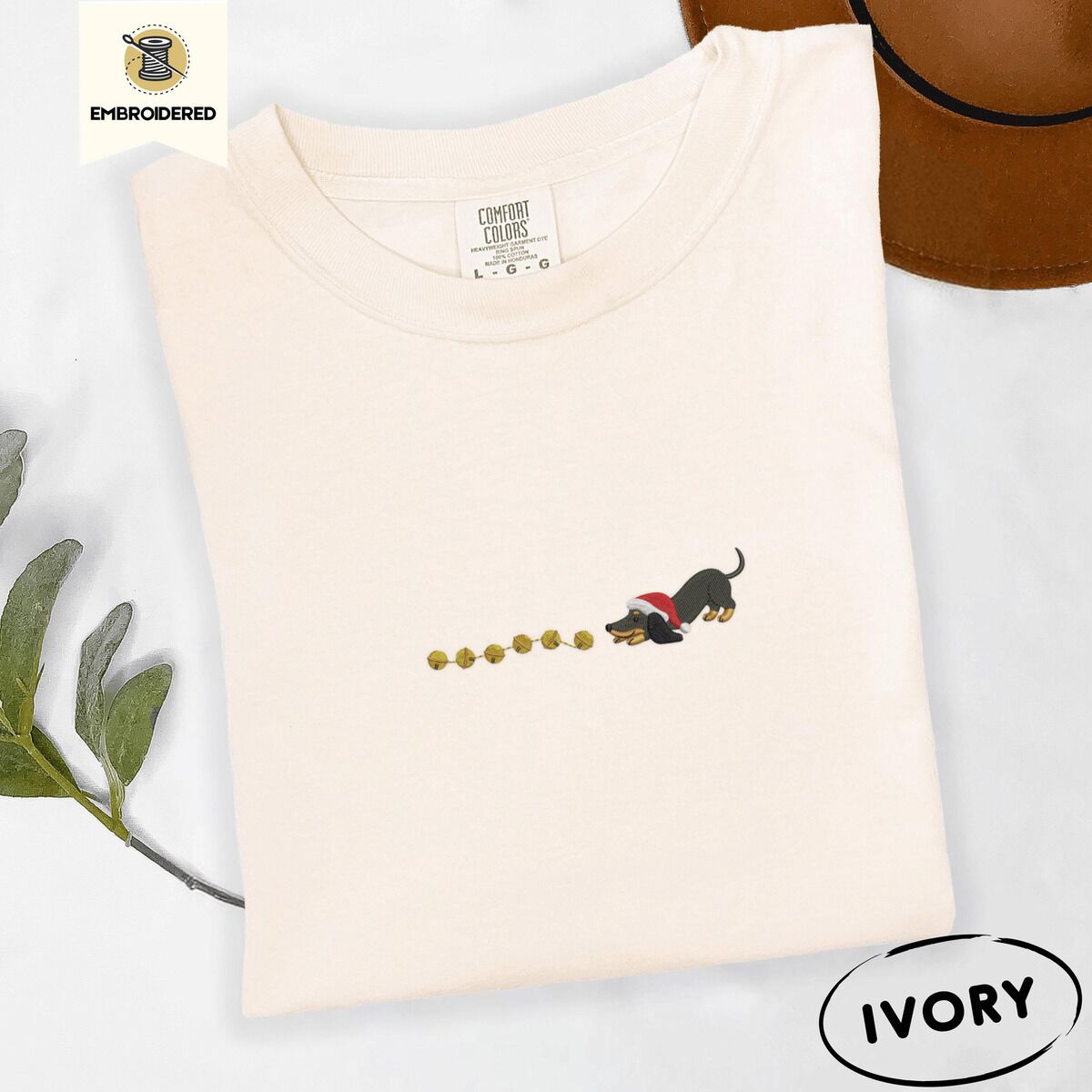 Dachshund Jingle Bells Shirt – Ivory Ivory Comfort Colors T-shirt embroidered with a black and tan dachshund in a Santa hat bowing down to sniff a string of gold jingle bells