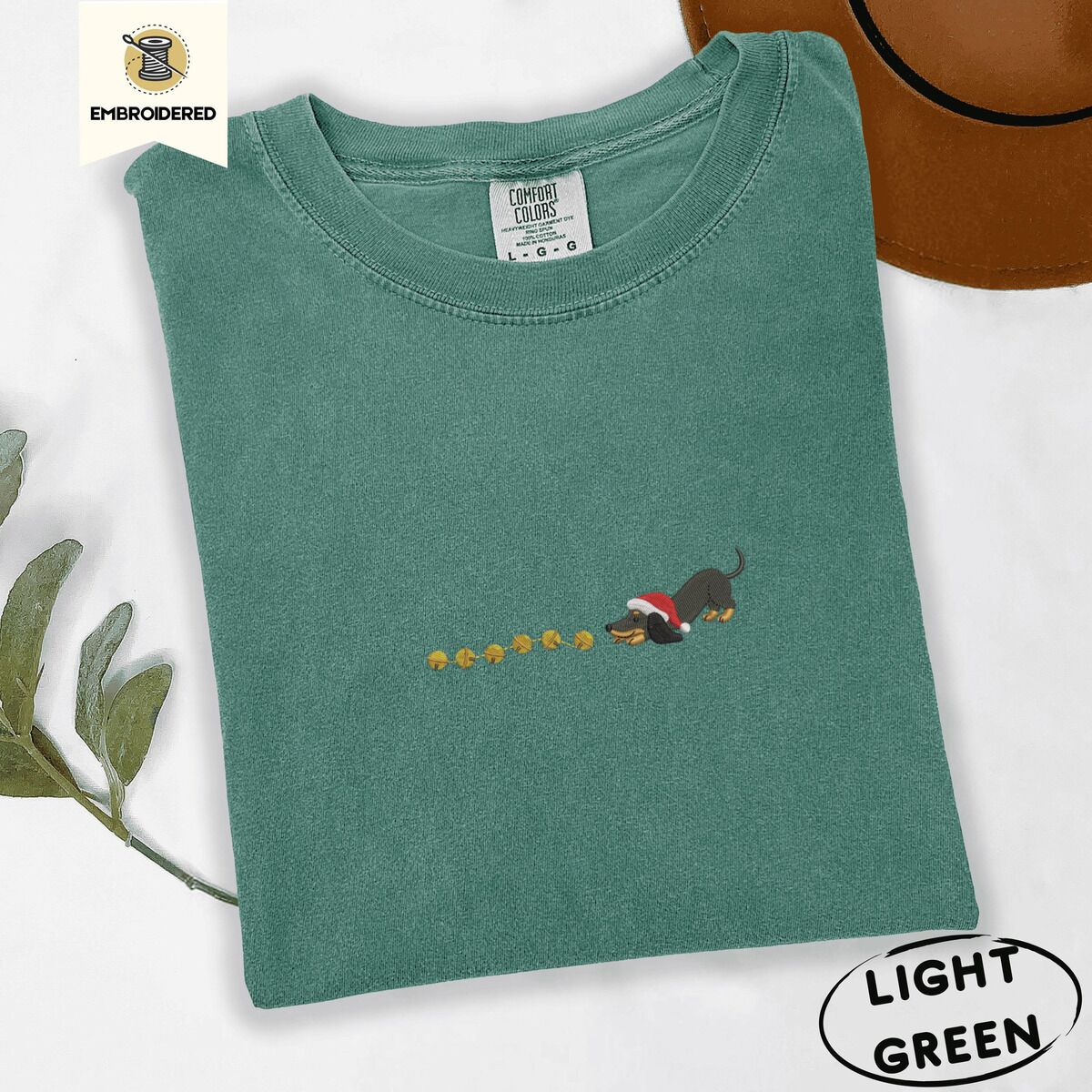 Dachshund Jingle Bells Shirt – Light Green Light Green Comfort Colors T-shirt embroidered with a black and tan dachshund in a Santa hat bowing down to sniff a string of gold jingle bells