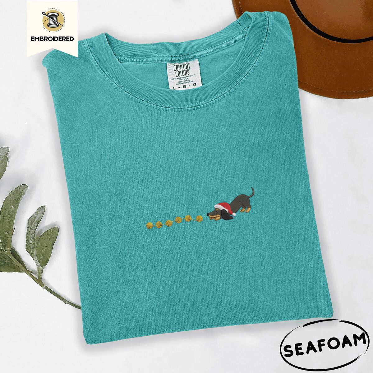 Dachshund Jingle Bells Shirt – Seafoam Seafoam Comfort Colors T-shirt embroidered with a black and tan dachshund in a Santa hat bowing down to sniff a string of gold jingle bells