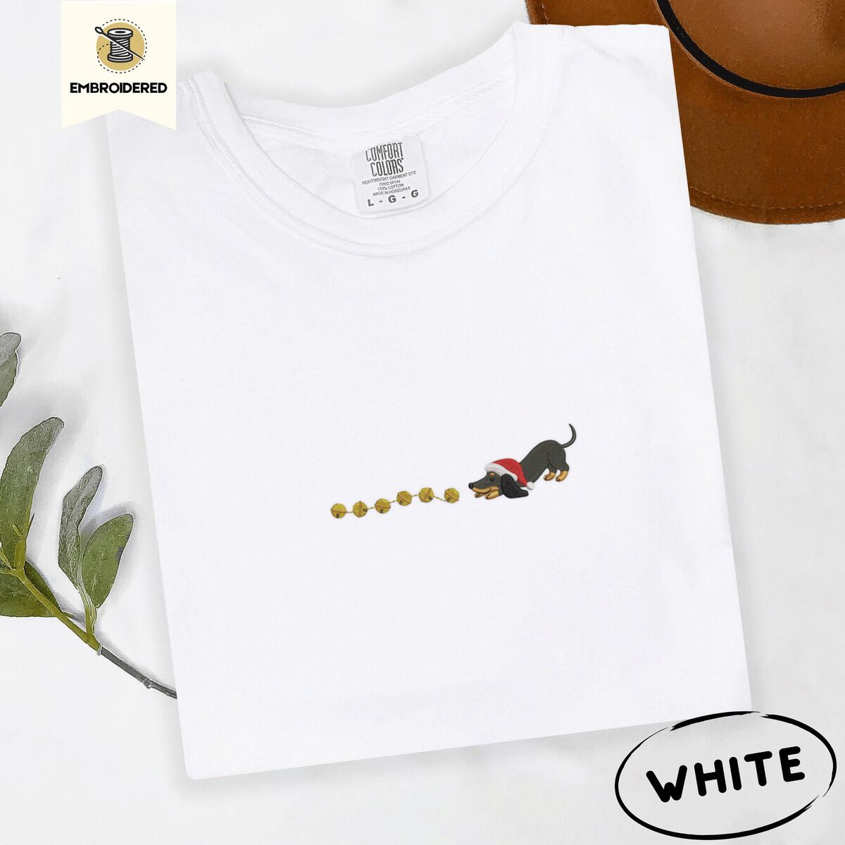 Dachshund Jingle Bells Shirt – White White Comfort Colors T-shirt embroidered with a black and tan dachshund in a Santa hat bowing down to sniff a string of gold jingle bells