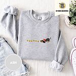 Ash crewneck sweatshirt folded with denim shorts, white sneakers and a potted plant, embroidered with a black and tan dachshund in a Santa hat bowing down to sniff a string of six gold jingle bells