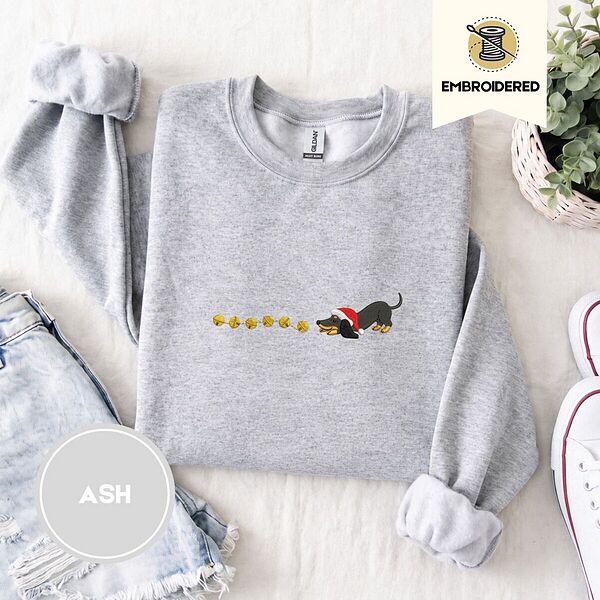 Ash crewneck sweatshirt folded with denim shorts, white sneakers and a potted plant, embroidered with a black and tan dachshund in a Santa hat bowing down to sniff a string of six gold jingle bells