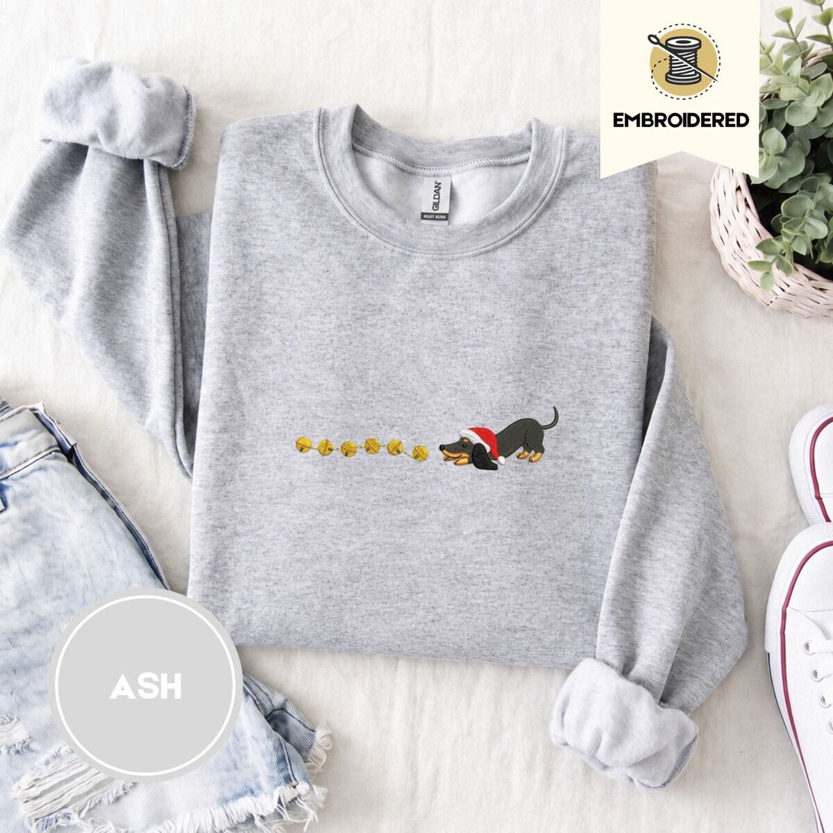 Dachshund Jingle Bells Sweatshirt – Ash Ash crewneck sweatshirt folded with denim shorts, white sneakers and a potted plant, embroidered with a black and tan dachshund in a Santa hat bowing down to sniff a string of six gold jingle bells