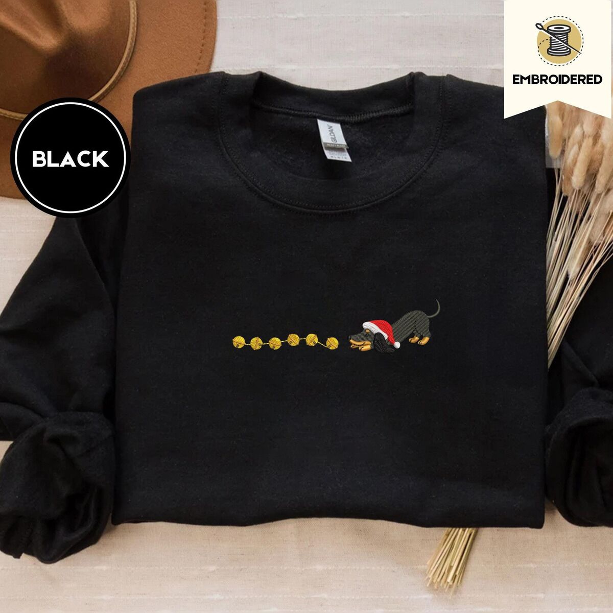 Dachshund Jingle Bells Sweatshirt – Black Black crewneck sweatshirt with a brown felt hat and dried bunny tail grass, embroidered with a black and tan dachshund in a Santa hat bowing down to sniff a string of six gold jingle bells
