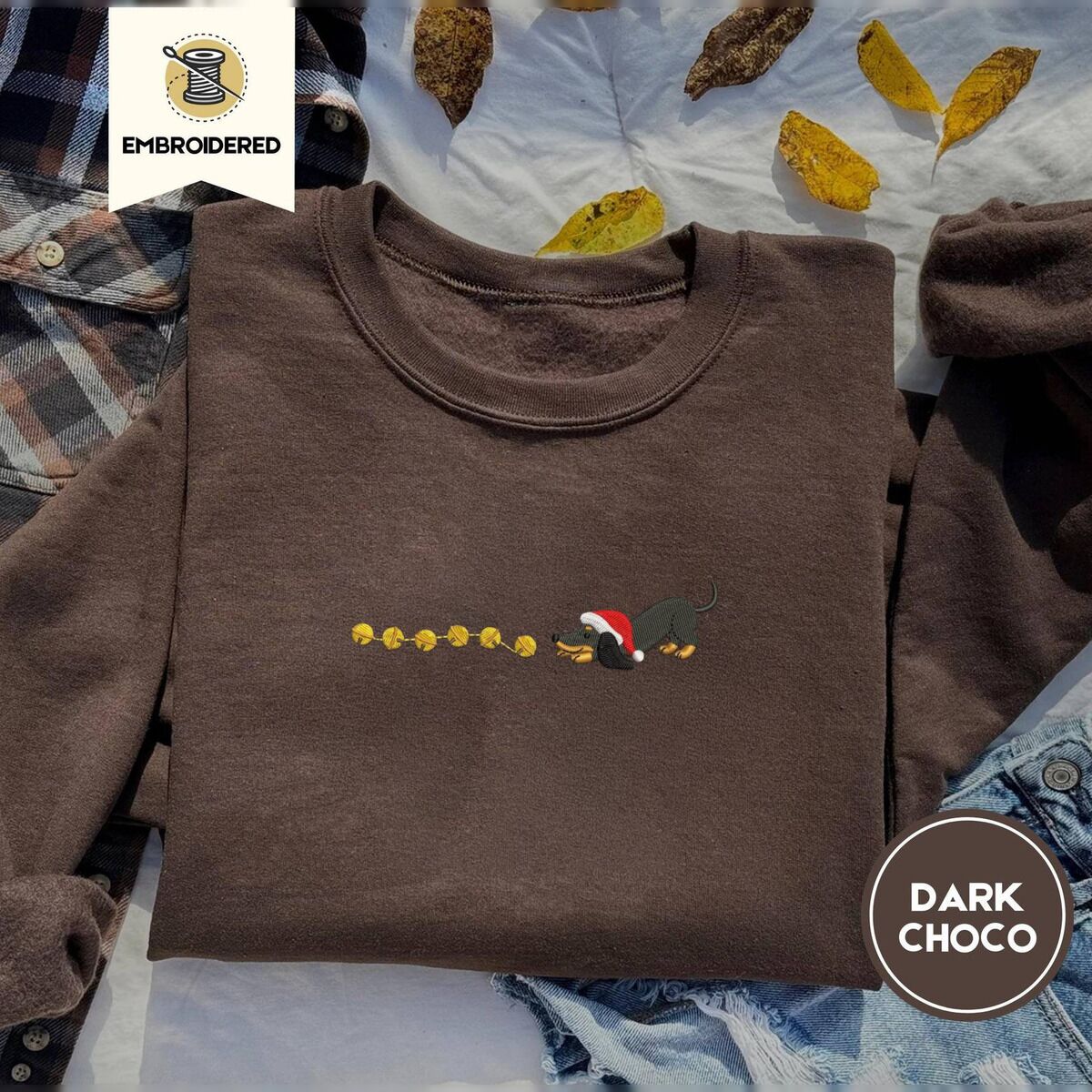 Dachshund Jingle Bells Sweatshirt – Dark Chocolate Dark Chocolate crewneck sweatshirt on a plaid flannel shirt with yellow autumn leaves and denim shorts, embroidered with a black and tan dachshund in a Santa hat bowing down to sniff a string of six gold jingle bells