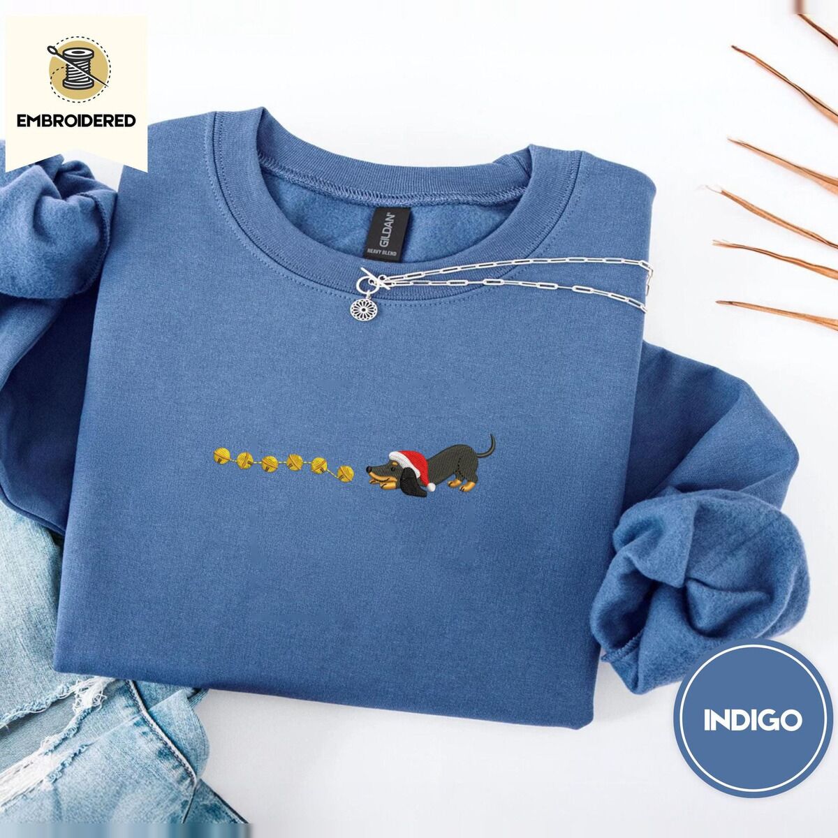Dachshund Jingle Bells Sweatshirt – Indigo Blue Indigo Blue crewneck sweatshirt folded with light jeans and dried twigs, embroidered with a black and tan dachshund in a Santa hat bowing down to sniff a string of six gold jingle bells