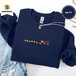 Navy crewneck sweatshirt folded with ripped jeans and a silver chain necklace, embroidered with a black and tan dachshund in a Santa hat bowing down to sniff a string of six gold jingle bells
