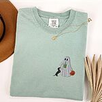 Bay T-shirt folded flat with a necklace, a brown hat and dried palm leaves, embroidered with a ghost sipping an iced matcha, a black and tan dachshund at her side and a jack-o'-lantern pail on the left chest