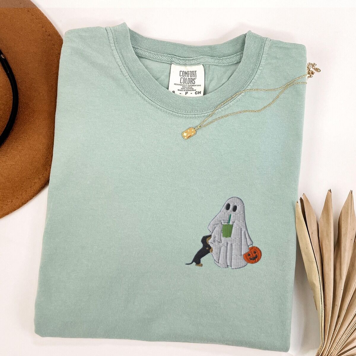 Dachshund Matcha Shirt – Bay Bay T-shirt folded flat with a necklace, a brown hat and dried palm leaves, embroidered with a ghost sipping an iced matcha, a black and tan dachshund at her side and a jack-o'-lantern pail on the left chest