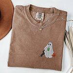 Espresso T-shirt folded flat with a necklace, a brown hat and dried palm leaves, embroidered with a ghost sipping an iced matcha, a black and tan dachshund at her side and a jack-o'-lantern pail on the left chest