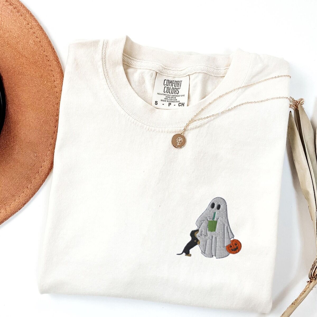 Dachshund Matcha Shirt – Ivory Ivory T-shirt folded flat with a necklace, a brown hat and dried palm leaves, embroidered with a ghost sipping an iced matcha, a black and tan dachshund at her side and a jack-o'-lantern pail on the left chest