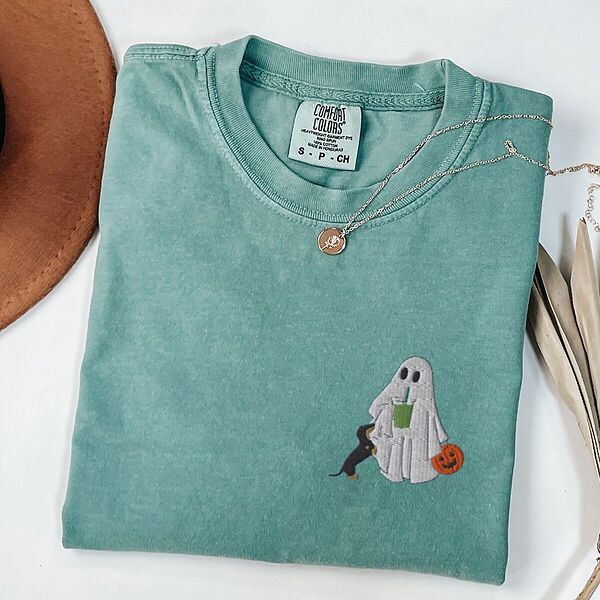 Light Green T-shirt folded flat with a necklace, a brown hat and dried palm leaves, embroidered with a ghost sipping an iced matcha, a black and tan dachshund at her side and a jack-o'-lantern pail on the left chest