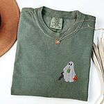 Moss T-shirt folded flat with a necklace, a brown hat and dried palm leaves, embroidered with a ghost sipping an iced matcha, a black and tan dachshund at her side and a jack-o'-lantern pail on the left chest