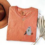 Terracotta T-shirt folded flat with a necklace, a brown hat and dried palm leaves, embroidered with a ghost sipping an iced matcha, a black and tan dachshund at her side and a jack-o'-lantern pail on the left chest