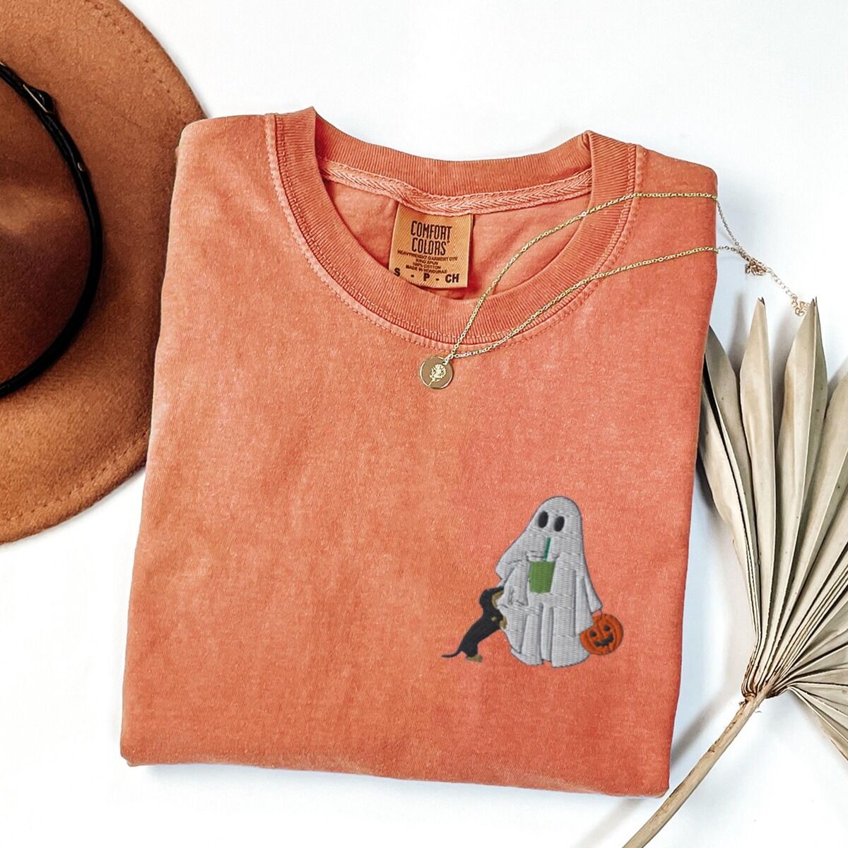 Dachshund Matcha Shirt – Terracotta Terracotta T-shirt folded flat with a necklace, a brown hat and dried palm leaves, embroidered with a ghost sipping an iced matcha, a black and tan dachshund at her side and a jack-o'-lantern pail on the left chest