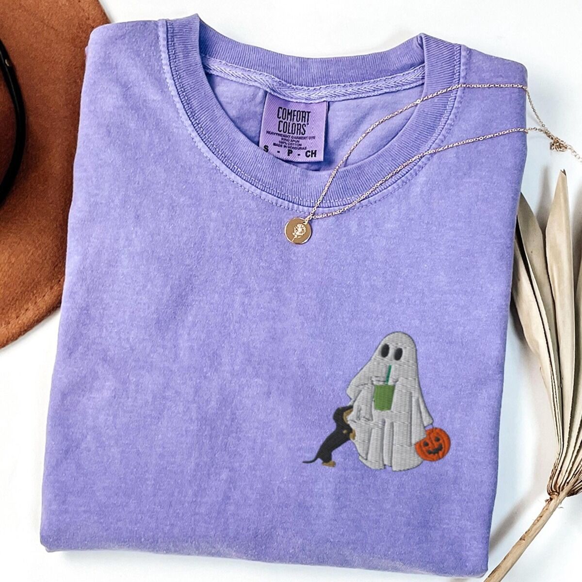 Dachshund Matcha Shirt – Violet Violet T-shirt folded flat with a necklace, a brown hat and dried palm leaves, embroidered with a ghost sipping an iced matcha, a black and tan dachshund at her side and a jack-o'-lantern pail on the left chest
