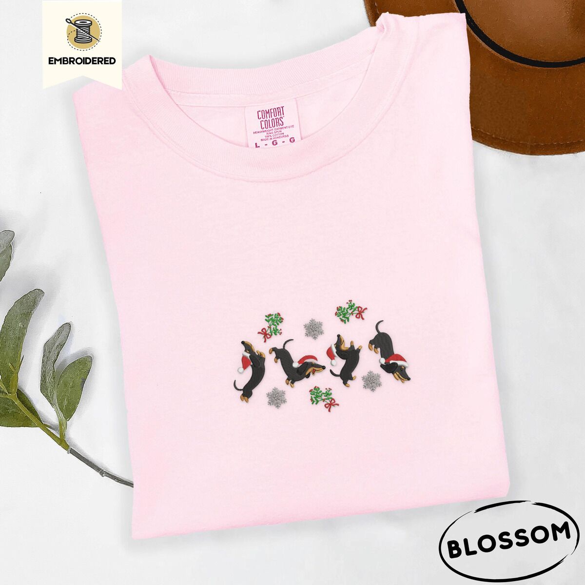 Dachshund Mistletoe Shirt – Blossom Blossom Comfort Colors T-shirt embroidered with four black and tan dachshunds in Santa hats leaping between sprigs of mistletoe and silver snowflakes