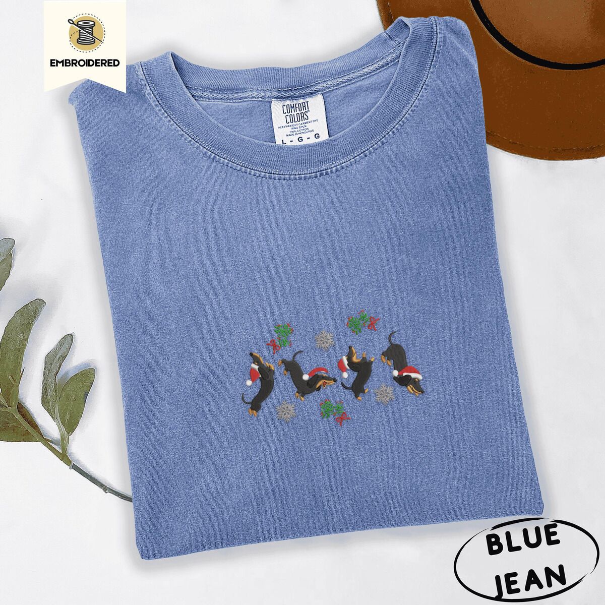 Dachshund Mistletoe Shirt – Blue Jean Blue Jean Comfort Colors T-shirt embroidered with four black and tan dachshunds in Santa hats leaping between sprigs of mistletoe and silver snowflakes