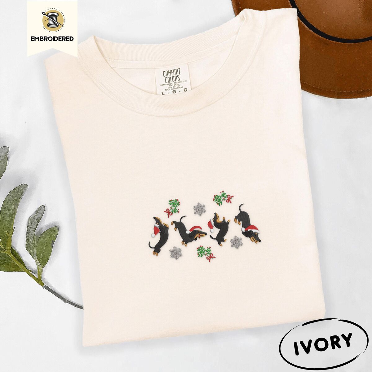 Dachshund Mistletoe Shirt – Ivory Ivory Comfort Colors T-shirt embroidered with four black and tan dachshunds in Santa hats leaping between sprigs of mistletoe and silver snowflakes