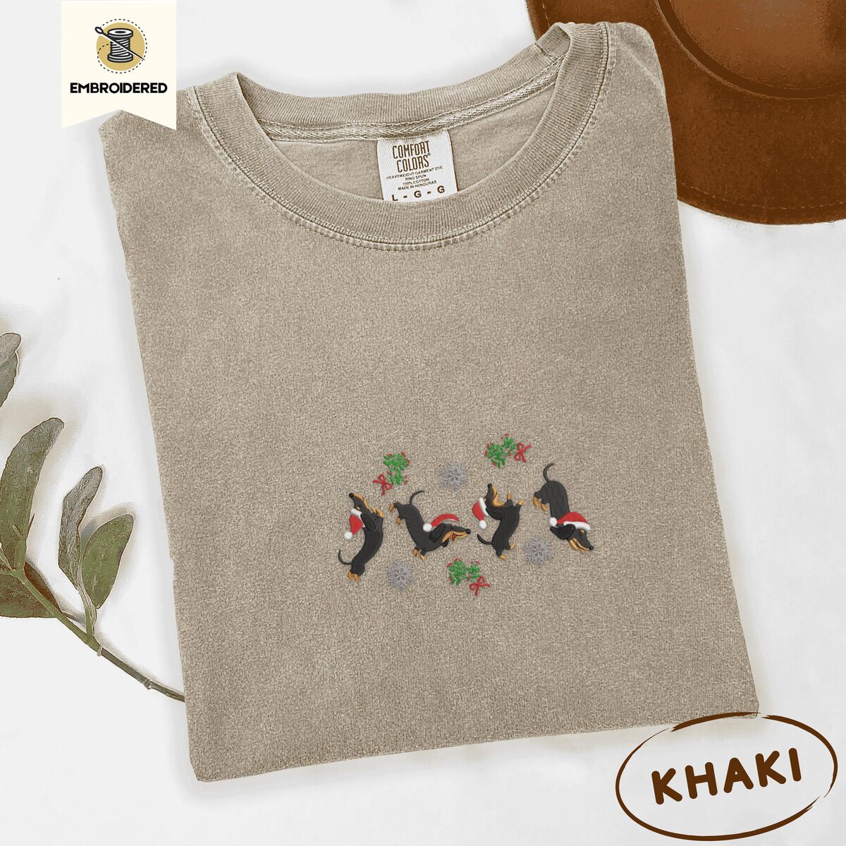 Dachshund Mistletoe Shirt – Khaki Khaki Comfort Colors T-shirt embroidered with four black and tan dachshunds in Santa hats leaping between sprigs of mistletoe and silver snowflakes