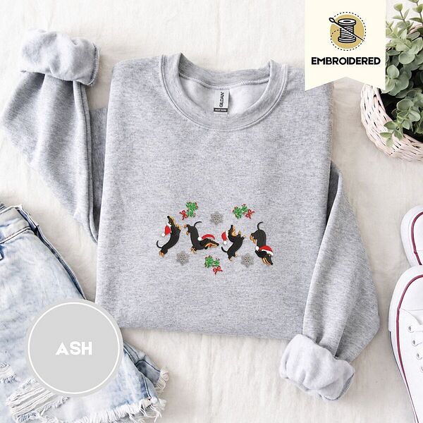 Ash crewneck sweatshirt folded with denim shorts, white sneakers and a potted plant, embroidered with four black and tan dachshunds in Santa hats leaping between mistletoe sprigs with red bows and silver snowflakes