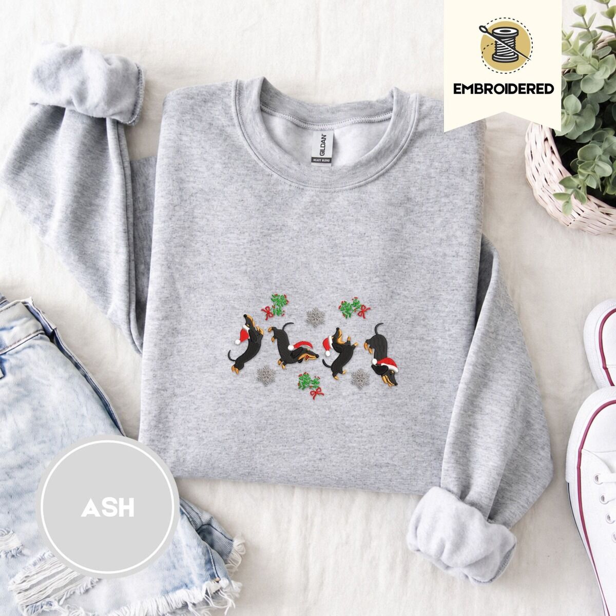 Dachshund Mistletoe Sweatshirt – Ash Ash crewneck sweatshirt folded with denim shorts, white sneakers and a potted plant, embroidered with four black and tan dachshunds in Santa hats leaping between mistletoe sprigs with red bows and silver snowflakes