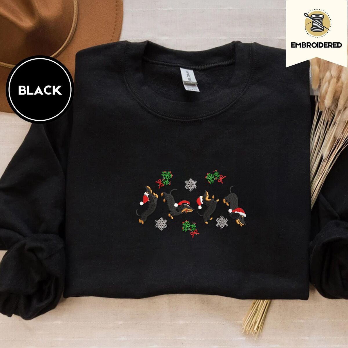 Dachshund Mistletoe Sweatshirt – Black Black crewneck sweatshirt with a brown felt hat and dried bunny tail grass, embroidered with four black and tan dachshunds in Santa hats leaping between mistletoe sprigs with red bows and silver snowflakes