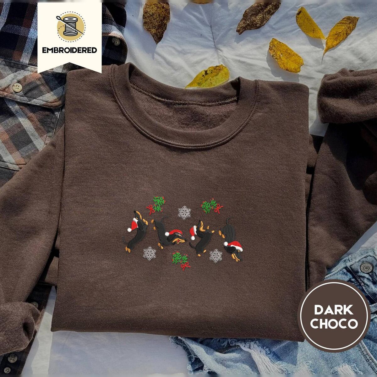 Dachshund Mistletoe Sweatshirt – Dark Chocolate Dark Chocolate crewneck sweatshirt on a plaid flannel shirt with yellow autumn leaves and denim shorts, embroidered with four black and tan dachshunds in Santa hats leaping between mistletoe sprigs with red bows and silver snowflakes