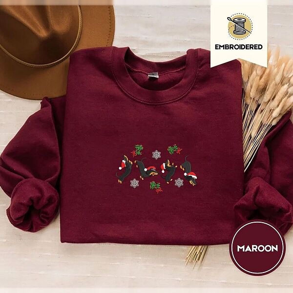 Maroon crewneck sweatshirt with a brown felt hat and dried bunny tail grass, embroidered with four black and tan dachshunds in Santa hats leaping between mistletoe sprigs with red bows and silver snowflakes