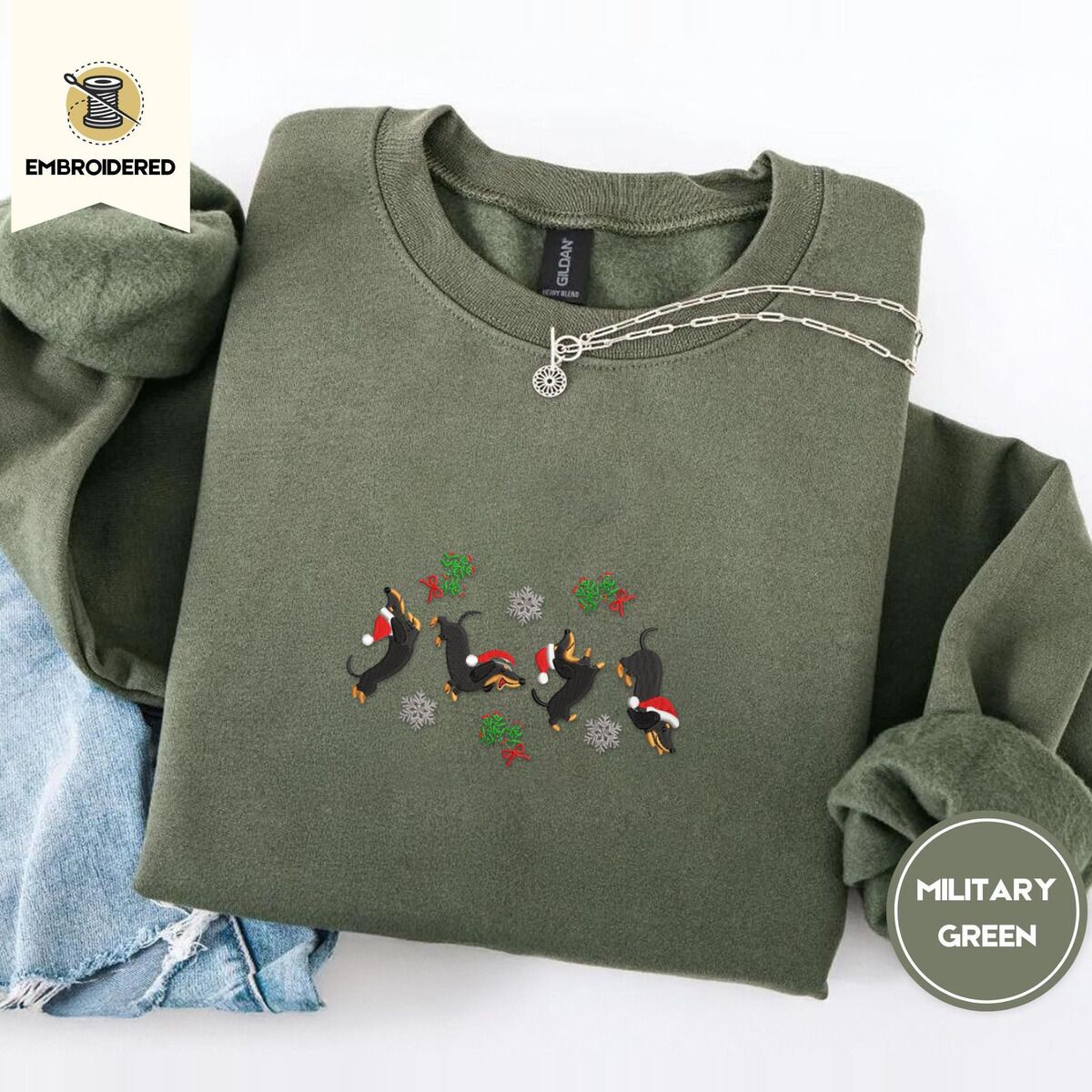 Dachshund Mistletoe Sweatshirt – Military Green Military Green crewneck sweatshirt folded with light jeans and a silver chain necklace, embroidered with four black and tan dachshunds in Santa hats leaping between mistletoe sprigs with red bows and silver snowflakes