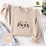 Sand crewneck sweatshirt folded with denim shorts, white sneakers and a potted plant, embroidered with four black and tan dachshunds in Santa hats leaping between mistletoe sprigs with red bows and silver snowflakes