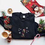 Black crewneck sweatshirt laid flat beside a Christmas stocking, a mug of coffee and a candy cane, embroidered with a sitting black and tan dachshund in a Santa hat and striped red scarf among white snowflakes