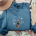 Indigo Blue crewneck sweatshirt laid flat with a felt hat and pampas grass, embroidered with a sitting black and tan dachshund in a Santa hat and striped red scarf among white snowflakes