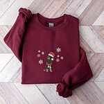 Maroon crewneck sweatshirt laid flat on a wood floor, embroidered with a sitting black and tan dachshund in a Santa hat and striped red scarf among white snowflakes