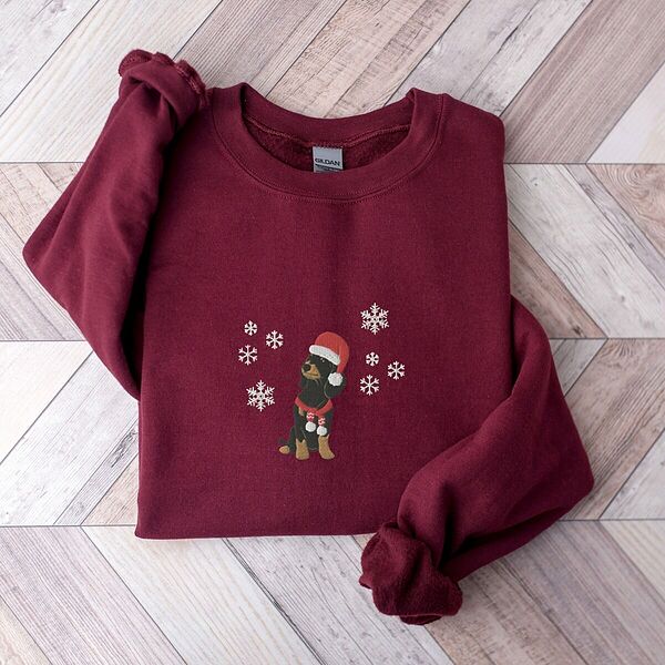 Maroon crewneck sweatshirt laid flat on a wood floor, embroidered with a sitting black and tan dachshund in a Santa hat and striped red scarf among white snowflakes