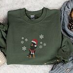 Military Green crewneck sweatshirt laid flat on a fur rug beside a knit blanket and pinecones, embroidered with a sitting black and tan dachshund in a Santa hat and striped red scarf among white snowflakes