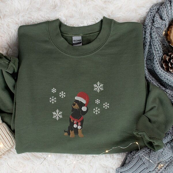Military Green crewneck sweatshirt laid flat on a fur rug beside a knit blanket and pinecones, embroidered with a sitting black and tan dachshund in a Santa hat and striped red scarf among white snowflakes
