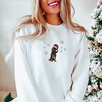 Smiling woman by a snowy Christmas tree in a White crewneck sweatshirt, embroidered with a sitting black and tan dachshund in a Santa hat and striped red scarf among white snowflakes