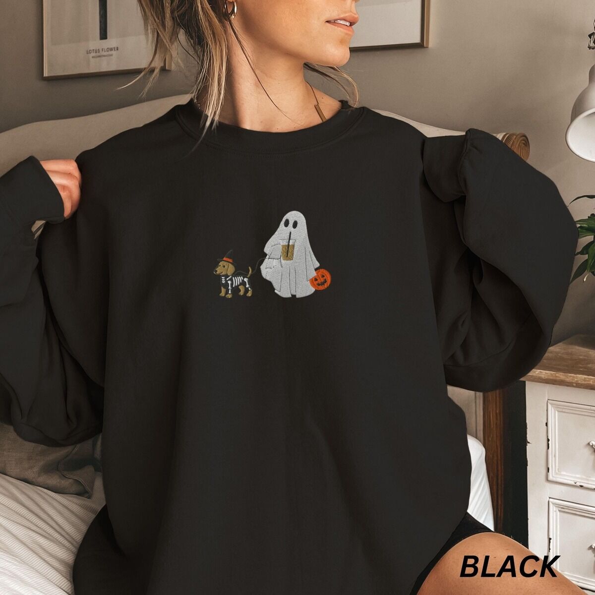 Dachshund Skeleton Sweatshirt – Black Woman in a bedroom wearing a Black crewneck sweatshirt embroidered with a red dachshund in a skeleton costume and witch hat on a leash held by a ghost sipping iced coffee