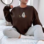 Woman sitting on a bed in a Dark Chocolate crewneck sweatshirt embroidered with a red dachshund in a skeleton costume and witch hat on a leash held by a ghost sipping iced coffee