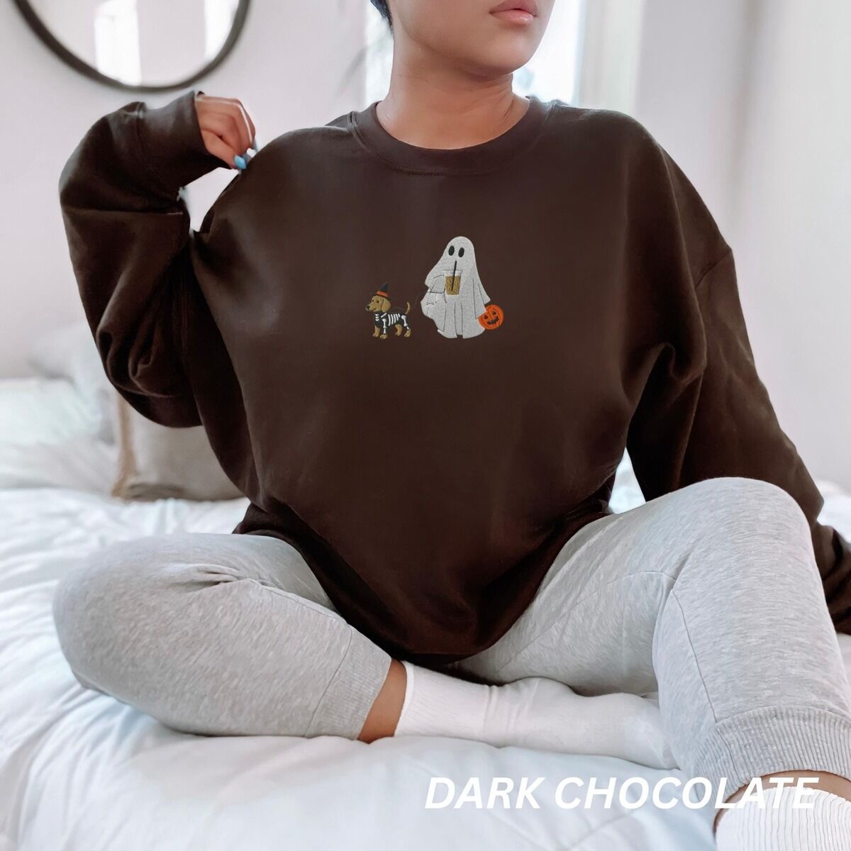 Dachshund Skeleton Sweatshirt – Dark Chocolate Woman sitting on a bed in a Dark Chocolate crewneck sweatshirt embroidered with a red dachshund in a skeleton costume and witch hat on a leash held by a ghost sipping iced coffee
