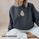 Woman sitting in a Dark Heather crewneck sweatshirt embroidered with a red dachshund in a skeleton costume and witch hat on a leash held by a ghost sipping iced coffee