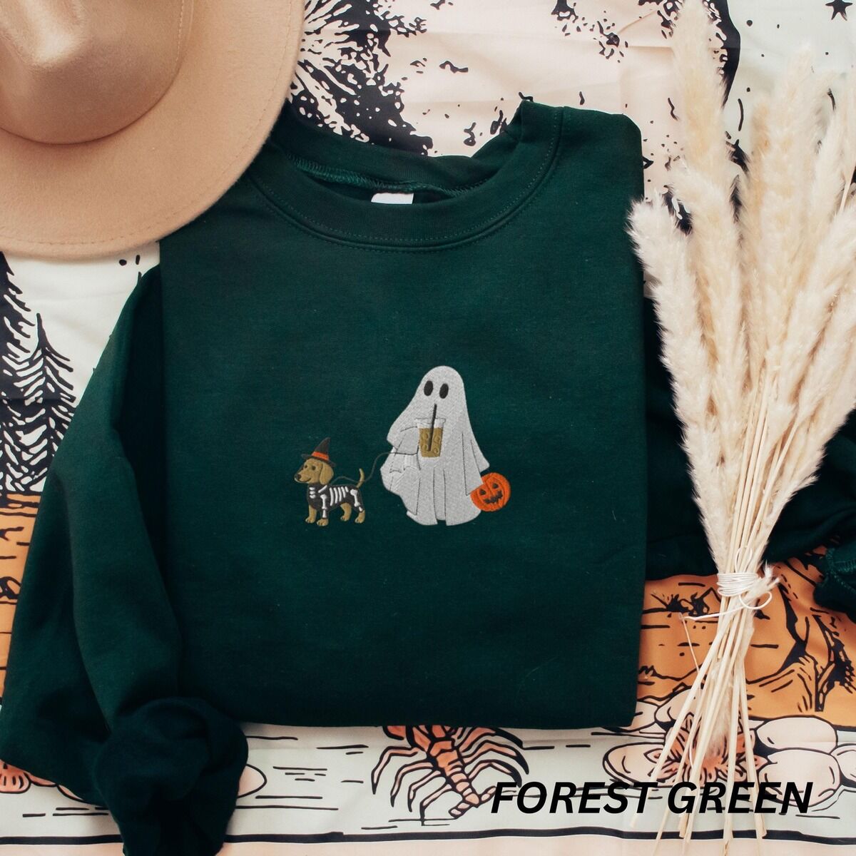 Dachshund Skeleton Sweatshirt – Forest Green Forest Green crewneck sweatshirt laid flat with a brown hat and pampas grass, embroidered with a red dachshund in a skeleton costume and witch hat on a leash held by a ghost sipping iced coffee